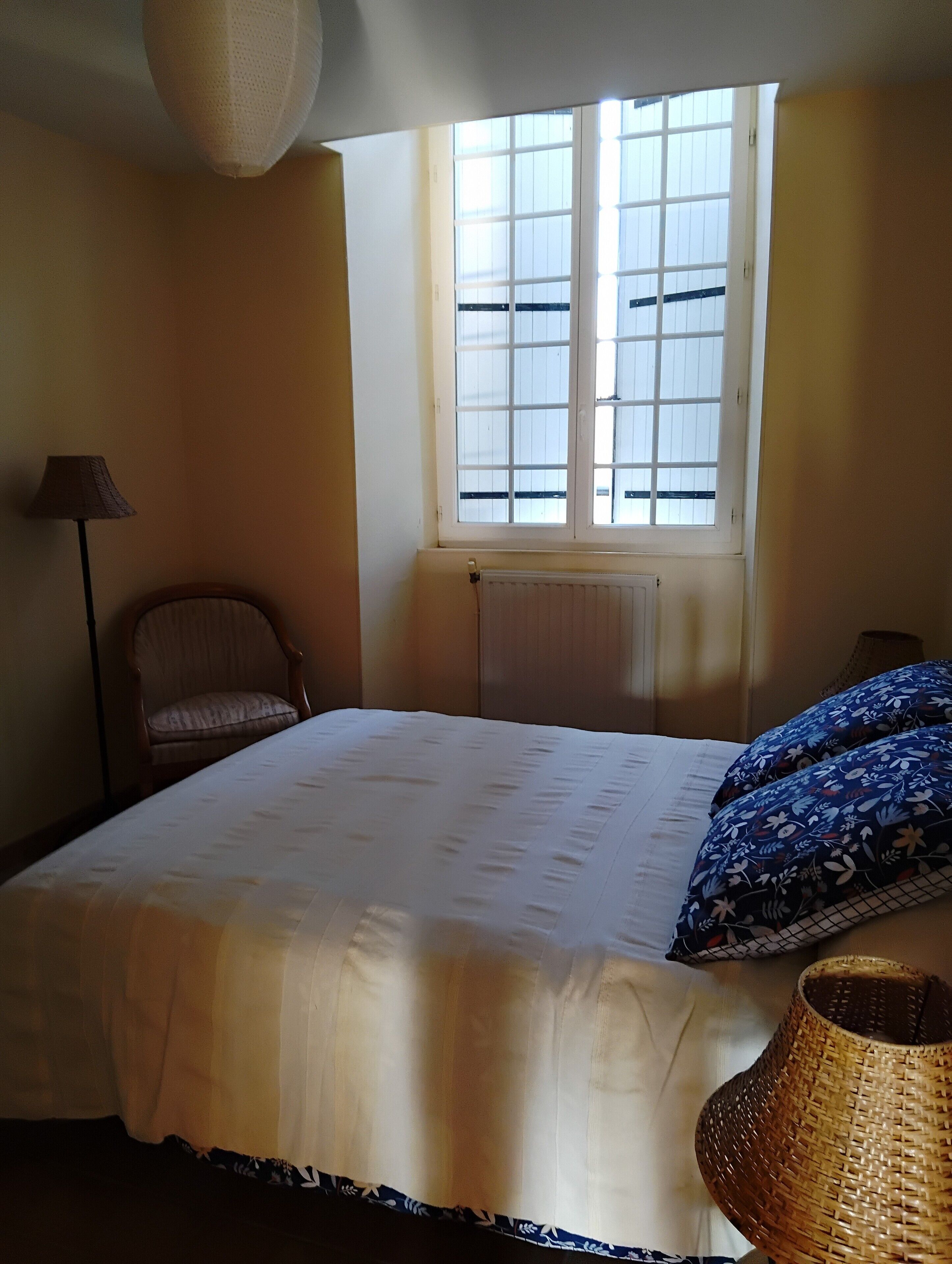 A bedroom with a bed, a chair, a lamp, a window, and a woven basket.