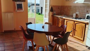 Un coin repas avec une table ronde et des chaises, une cuisine équipée d’une cuisinière et d’un évier, et une vue sur un jardin, accessible par une porte.