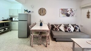 A compact living space with a kitchenette, a dining table with chairs, a sofa with decorative pillows, a wall clock, and a hanging light fixture. - 98 AN - Stay Heaven Apartment (Ayia Napa)