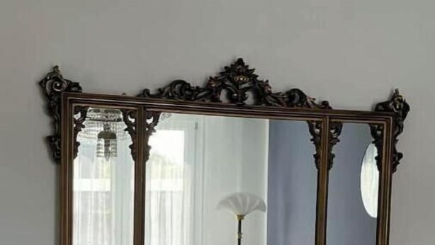 Ein großer, aufwendig verzierter Spiegel mit dunklem Rahmen, der ein Bett mit blauer Bettwäsche und eine Stehlampe reflektiert.