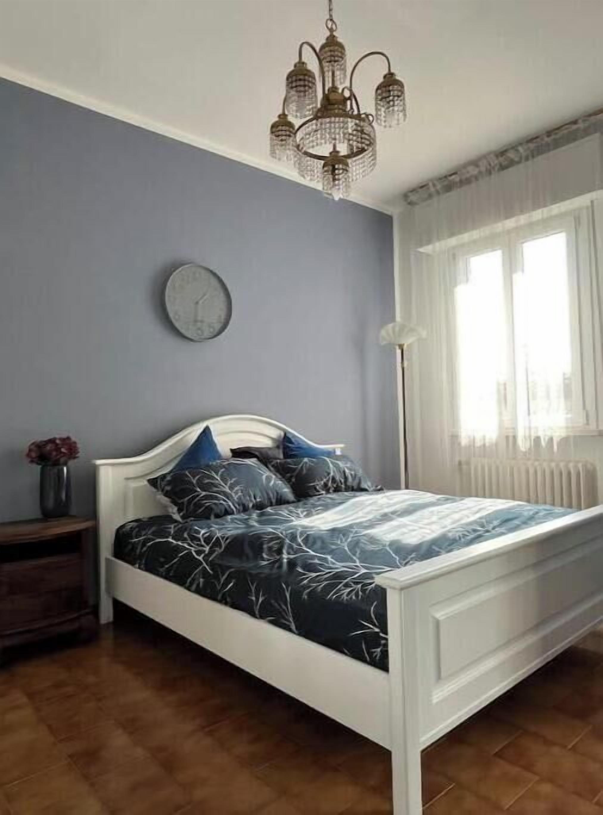 Un dormitorio con cama, un reloj, un radiador y una ventana con cortinas.