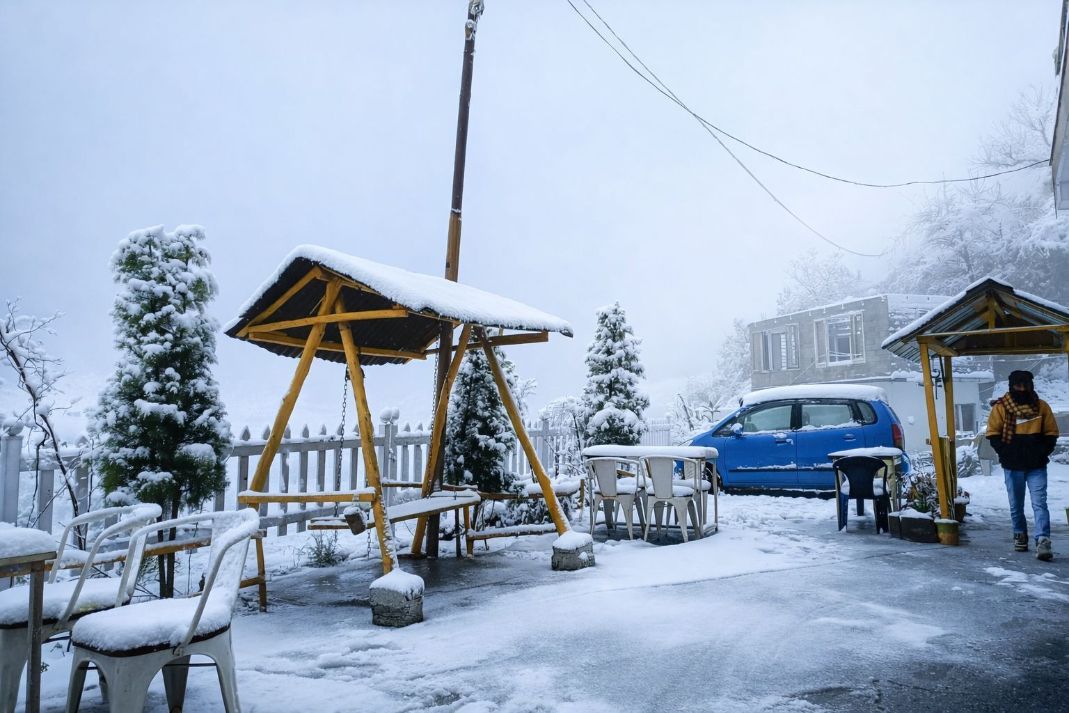 Un espace extérieur enneigé avec une balançoire, des tables, des chaises et une voiture bleue garée à proximité.