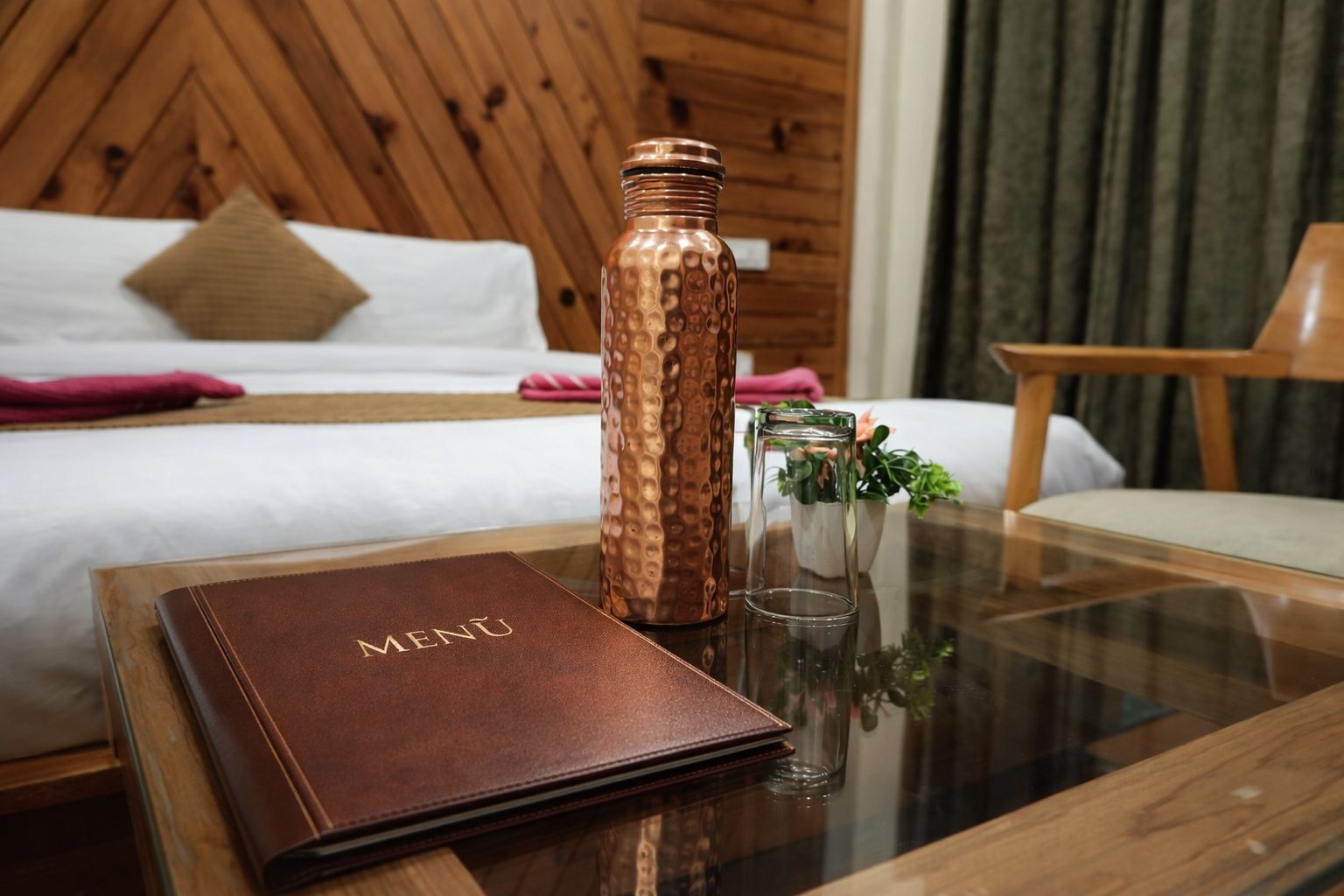 Ein ordentlich eingerichtetes Hotelzimmer mit einem Bett, einem Holz-Kopfteil, einem Glastisch mit einer Speisekarte, einer Kupfer-Trinkflasche und einer kleinen Topfpflanze.