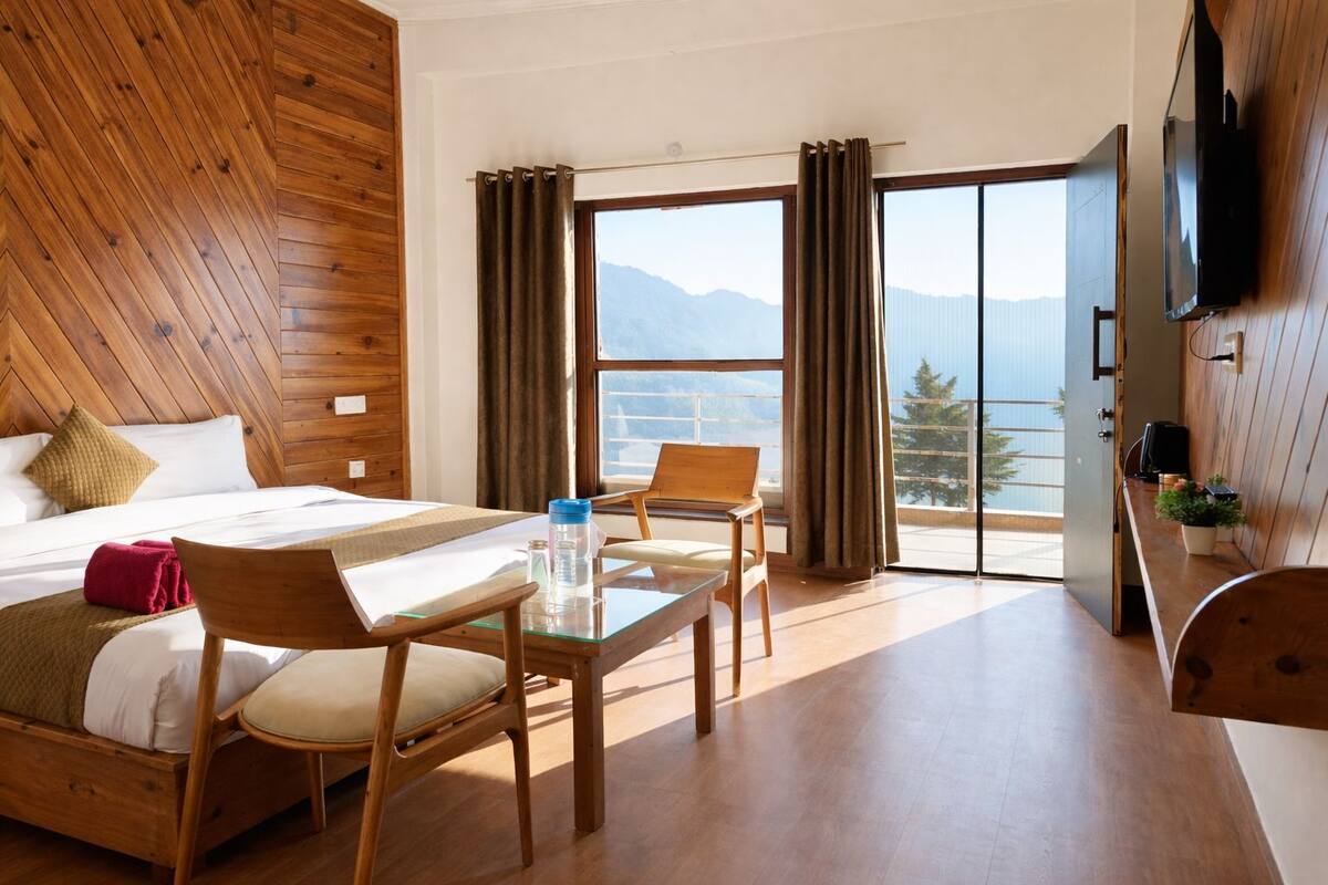 A hotel room with a bed, a table, chairs, a TV, and a view of mountains through the window.