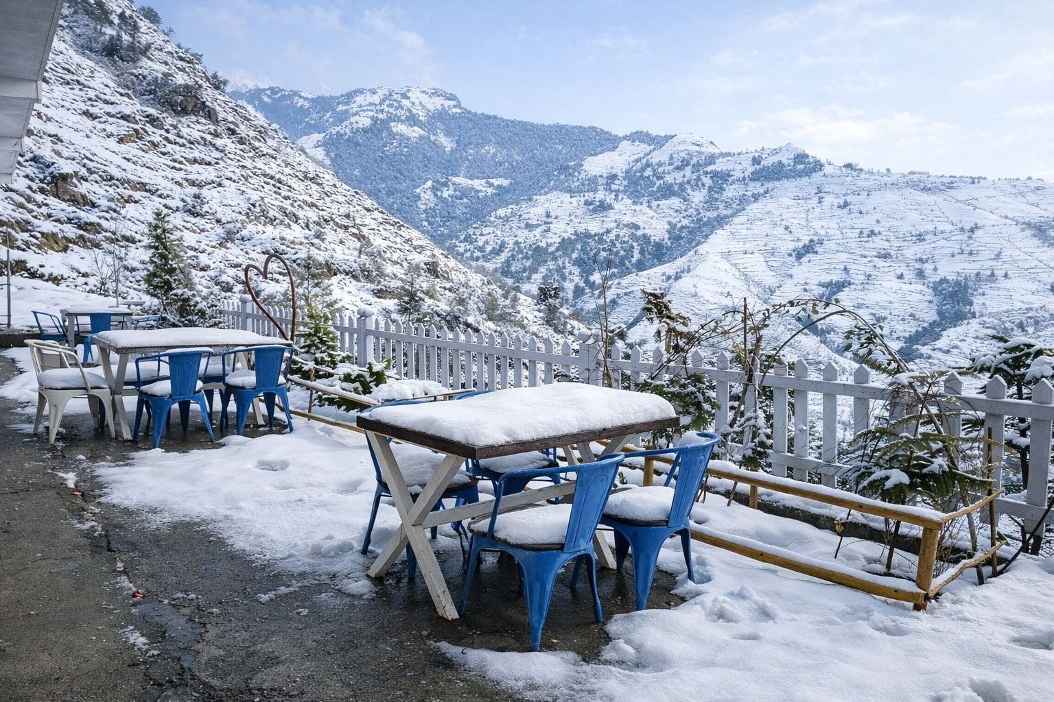 Mesas e cadeiras ao ar livre cobertas de neve, com montanhas ao fundo.