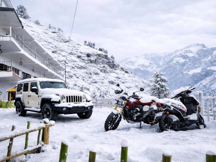 Zona exterior cubierta de nieve con un SUV blanco y dos motocicletas estacionadas cerca de un edificio con balcones.