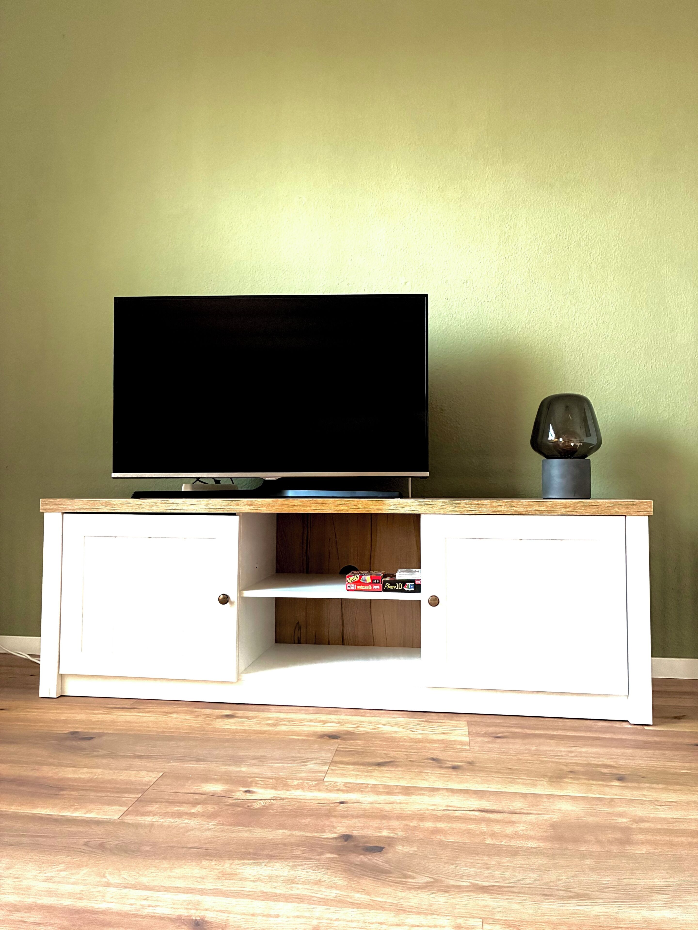 A TV on a wooden cabinet with a shelf below, a small decorative item, and a plain wall.