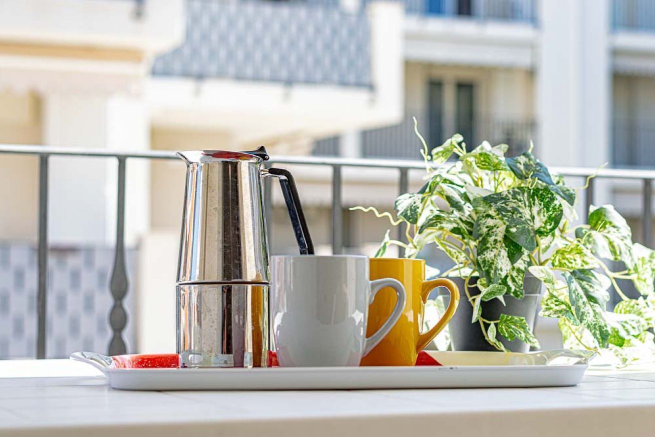 A balcony with a coffee maker, a white mug, a yellow mug, and a potted plant.