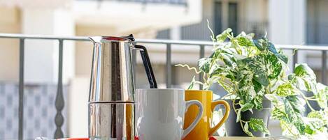 A balcony with a coffee maker, a white mug, a yellow mug, and a potted plant.