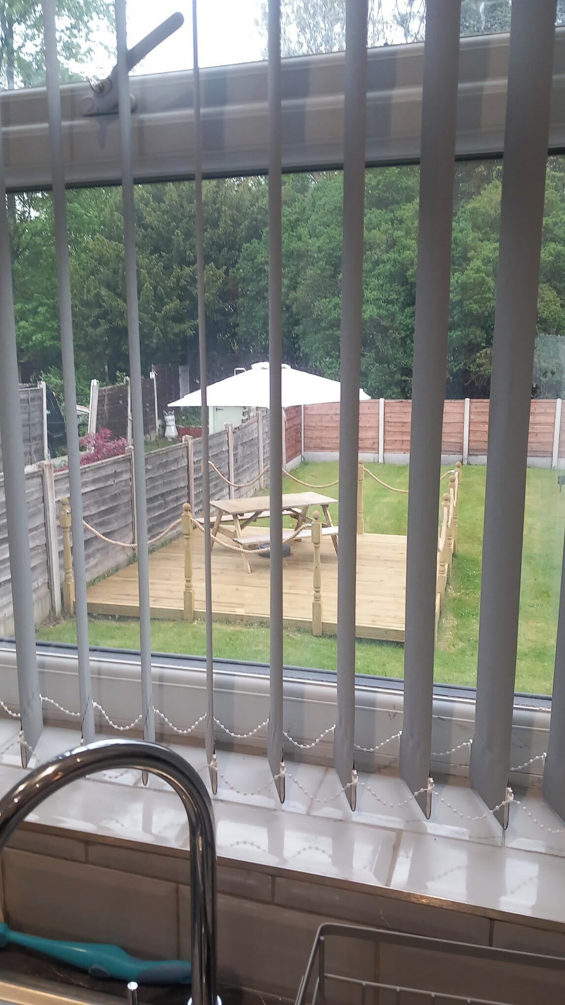 Une vue à travers une fenêtre avec des barreaux en métal, une terrasse en bois et une cour clôturée avec une structure blanche.