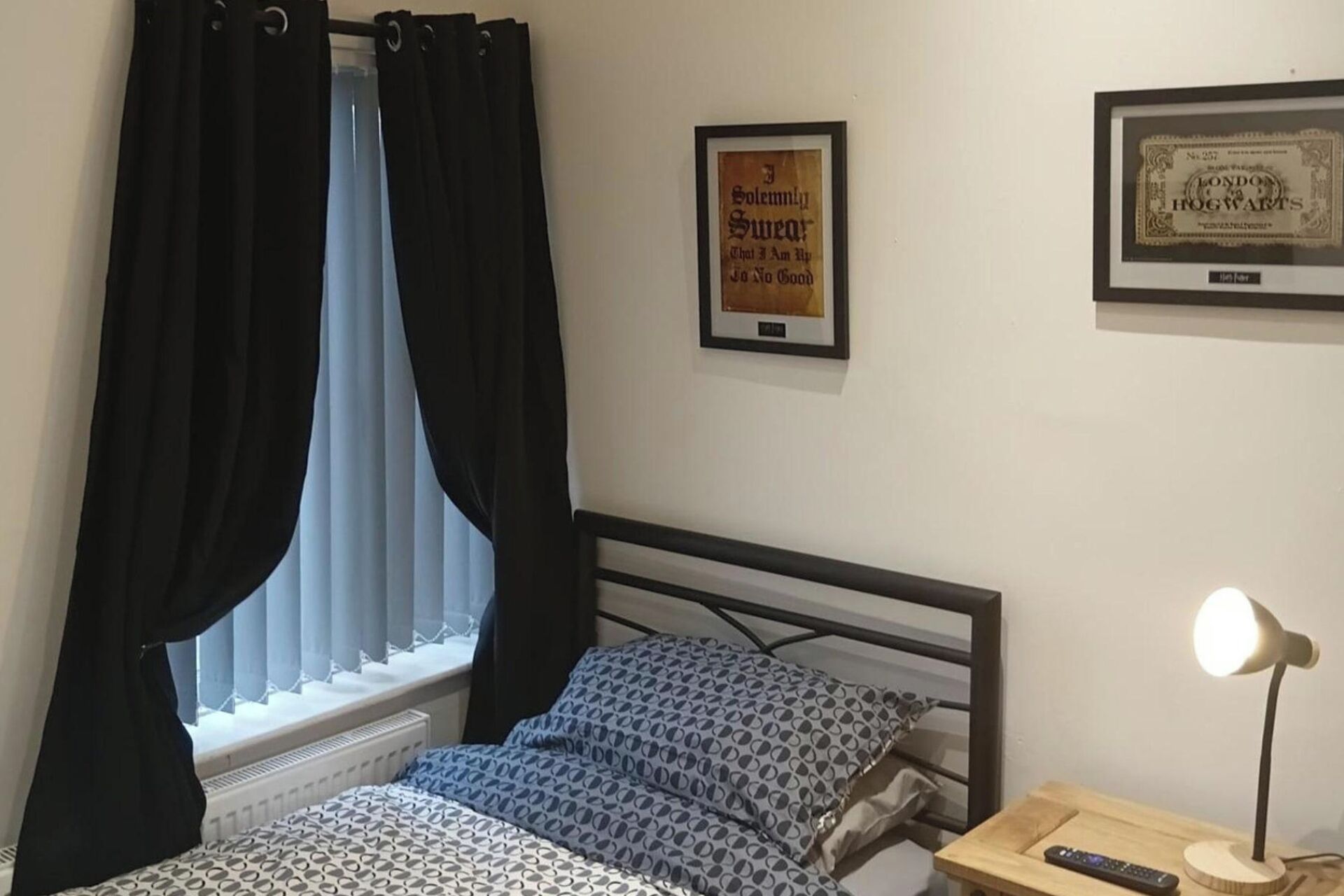 Une chambre à coucher avec un lit, une fenêtre avec des rideaux, une lampe de chevet et deux tableaux encadrés accrochés au mur.