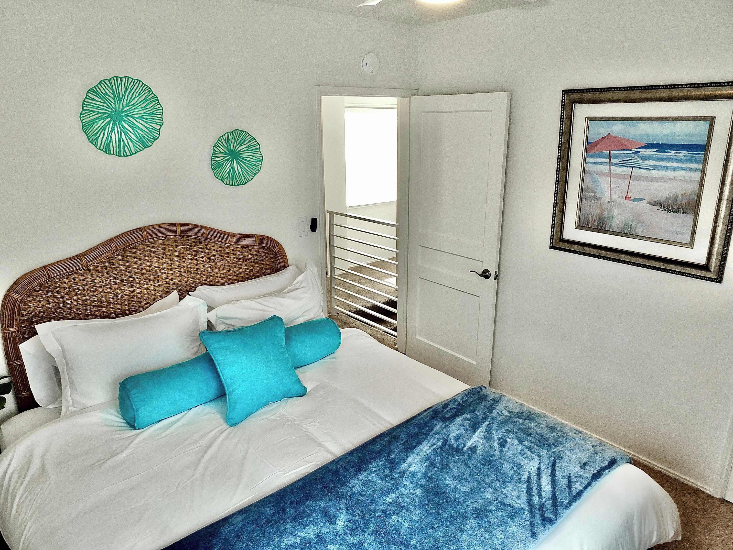 Een slaapkamer met een rieten hoofdbord, wit beddengoed, twee blauwe kussens, een blauwe deken en een schilderij van een strand in een lijst aan de muur.