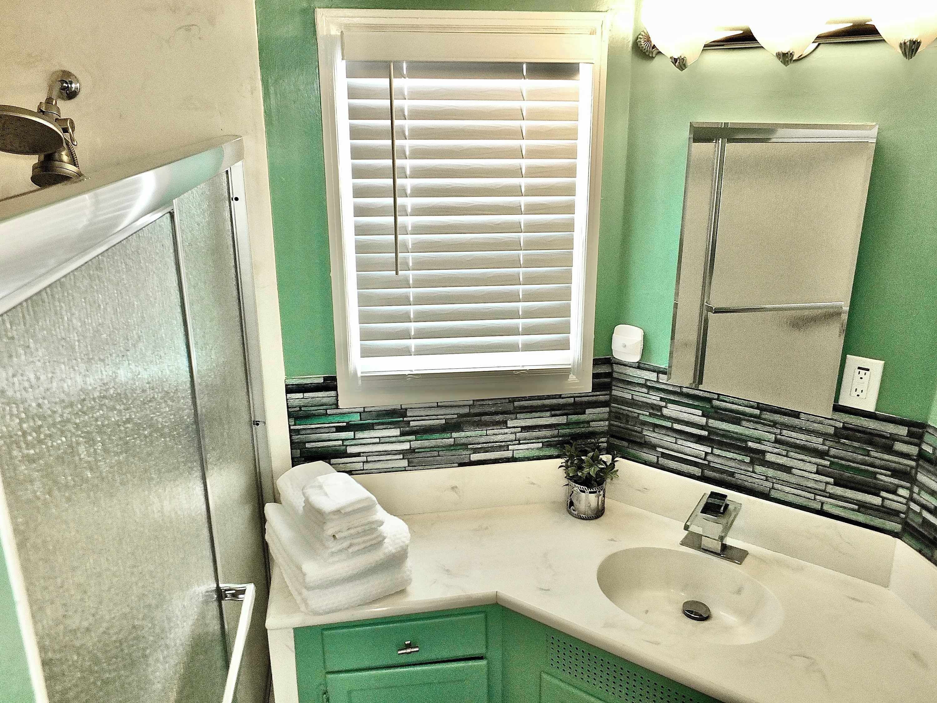 A bathroom with a white sink, green cabinets, a window with blinds, and a shower area.