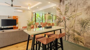 Una sala de estar moderna con televisor de pantalla plana, un sofá, una zona de comedor con mesa y taburetes de bar, y un gran mural tropical.