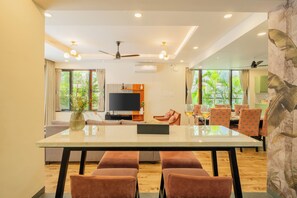 A modern living room with a large TV, a dining area with a glass table, and a ceiling fan. - ELIVAAS Villa Valerie 3BHK Pool & Bar (Siolim)
