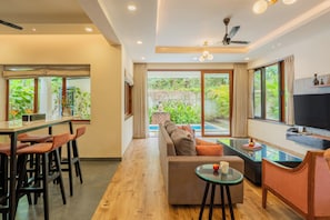A modern living room with a sofa, a dining area with chairs, a TV, and a view of greenery outside. - ELIVAAS Villa Valerie 3BHK Pool & Bar (Siolim)