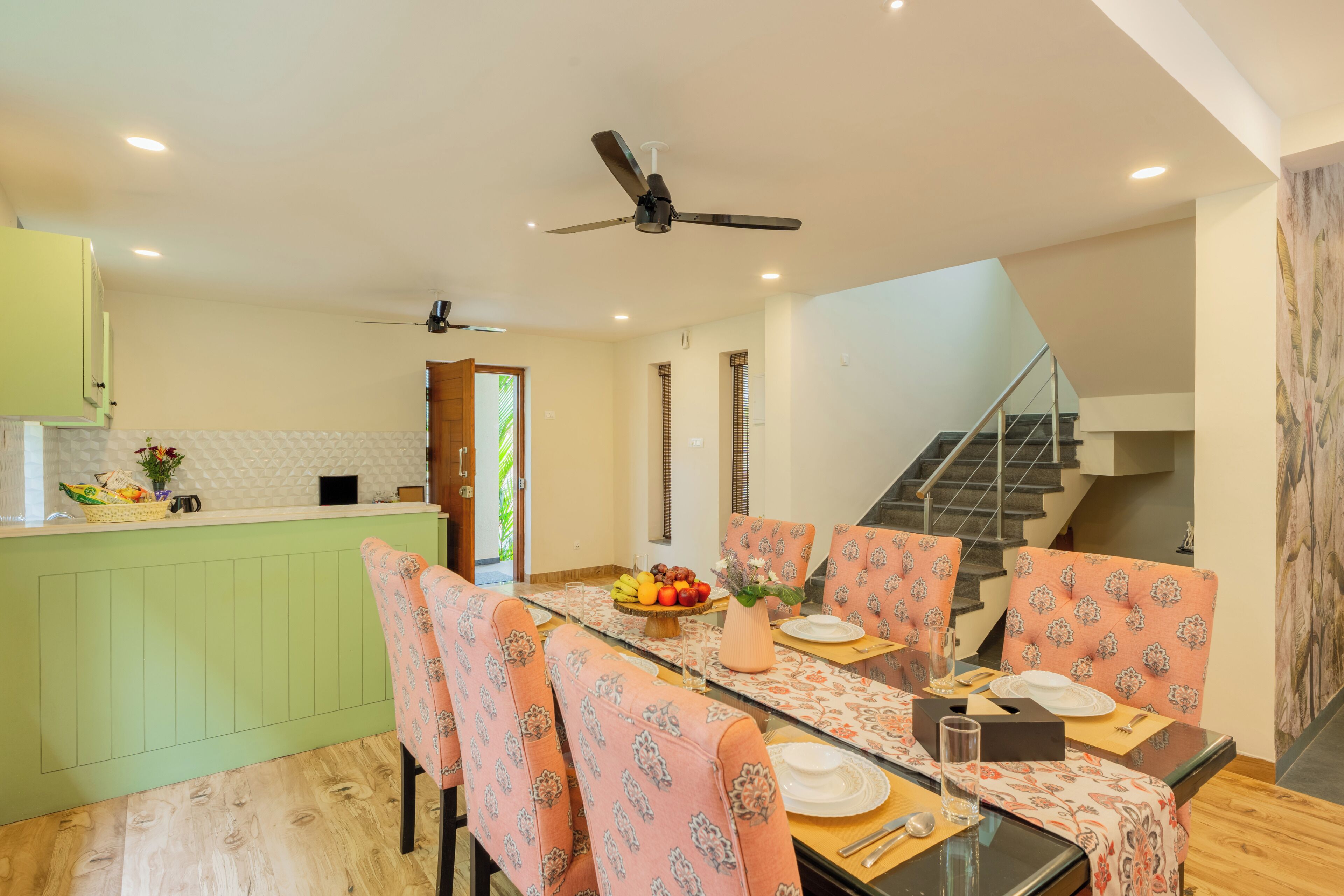 A dining area with a glass table, patterned chairs, a ceiling fan, and a staircase.