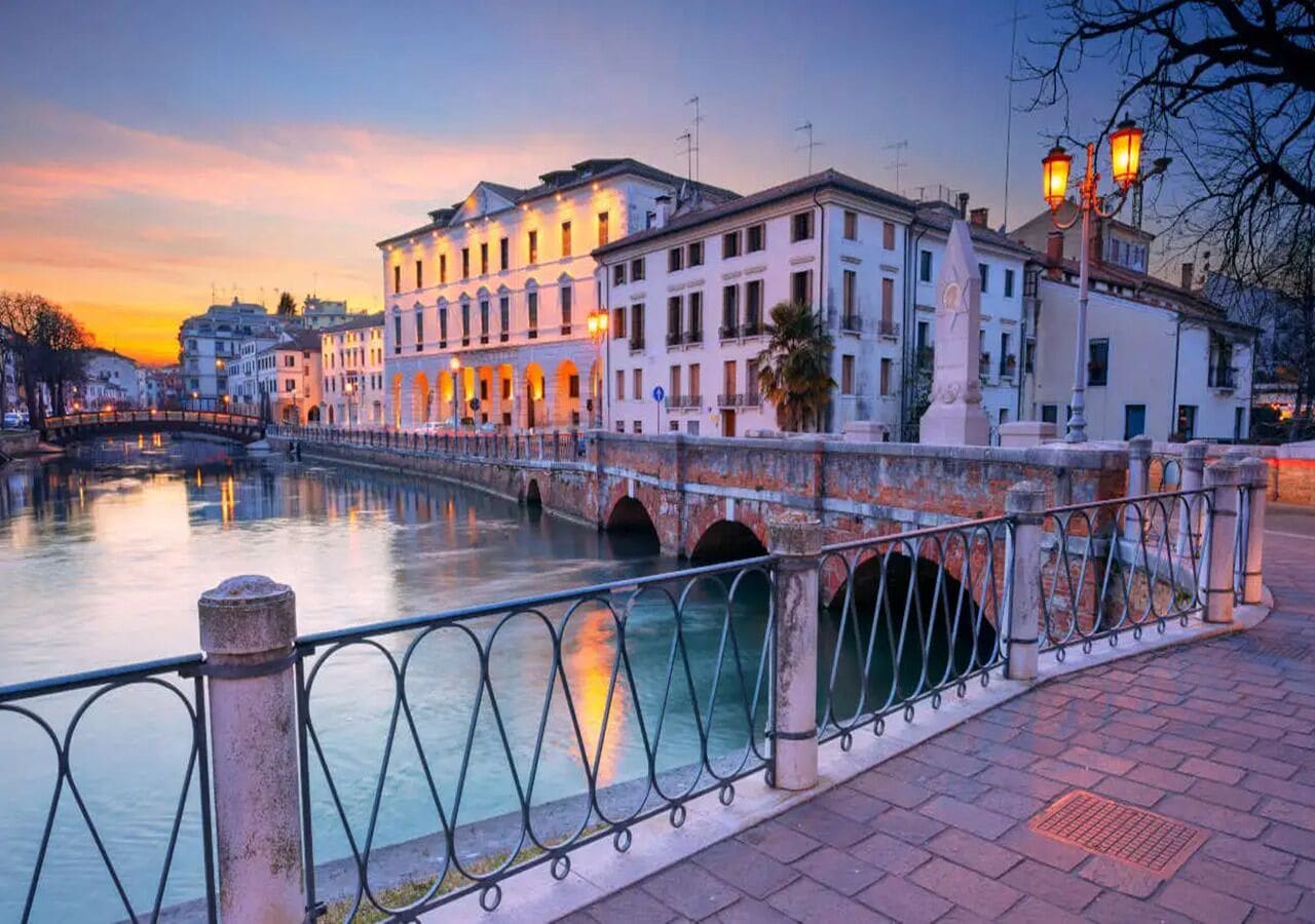 A riverside walkway with buildings, a bridge, and a railing.
