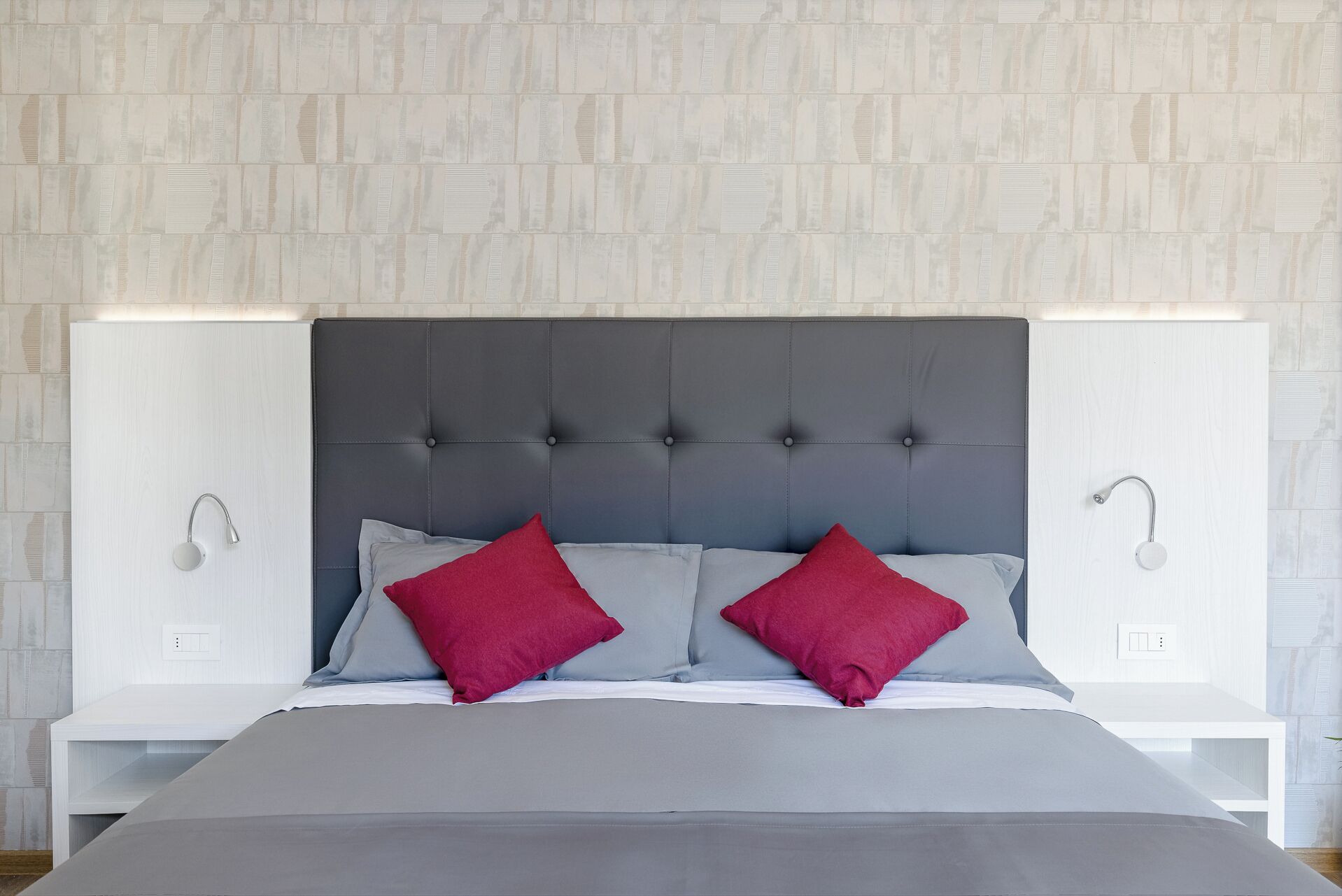 Una cama con cabecera tapizada de gris, dos almohadas rojas y una mesita de noche blanca con una lámpara.