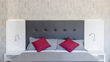 Una cama con cabecera tapizada de gris, dos almohadas rojas y una mesita de noche blanca con una lámpara.