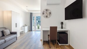 Une pièce moderne et bien éclairée, avec un plancher en bois, un canapé gris, un bureau avec une chaise, une horloge murale et un téléviseur.