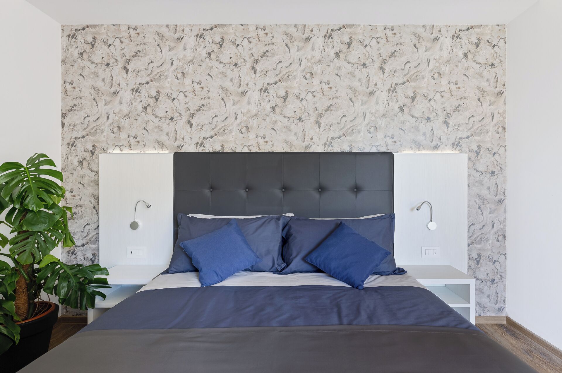 Camera da letto con parete a effetto marmo, testiera imbottita, due comodini e un'ampia pianta in vaso.