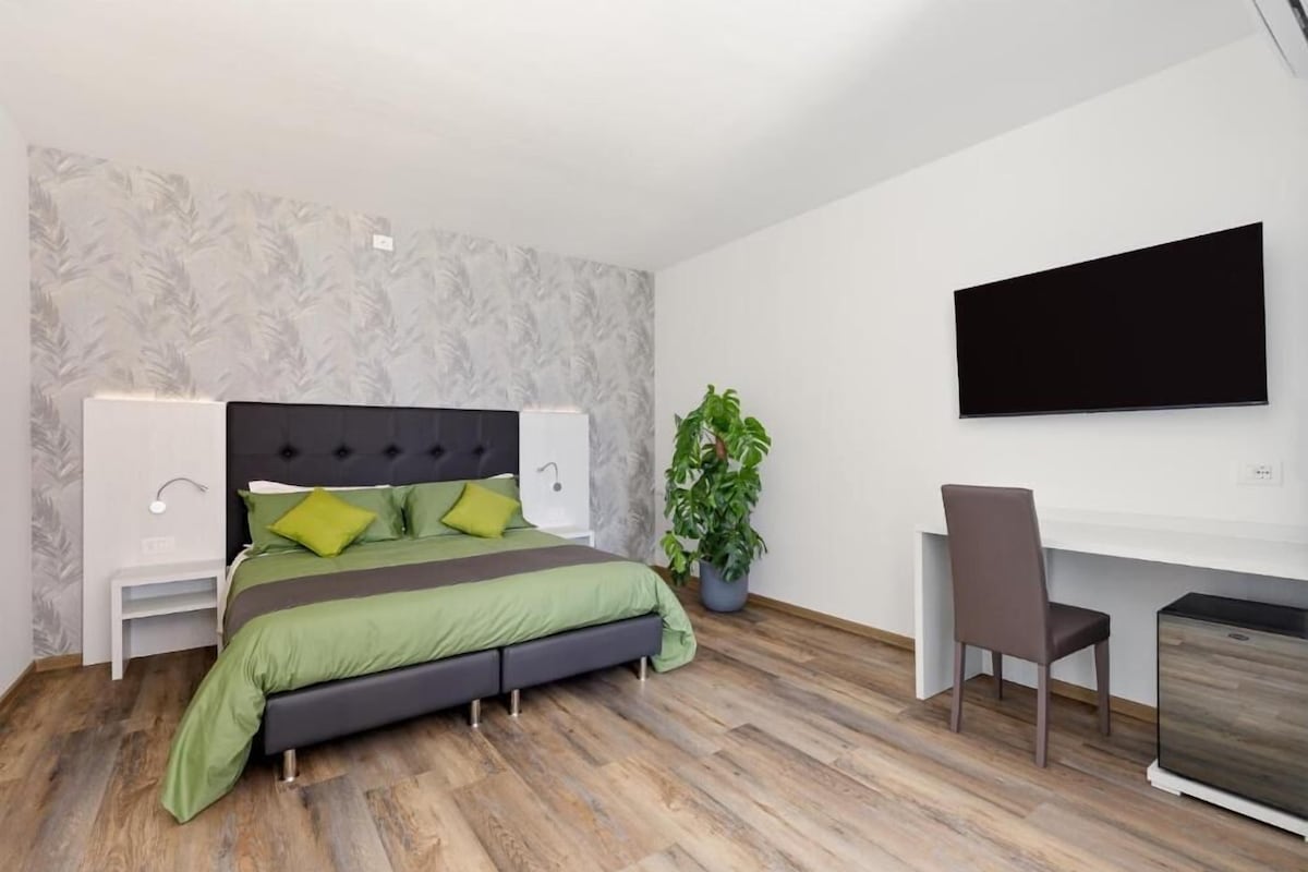 Un dormitorio con cama, mesitas de noche, un escritorio, una silla, una planta en maceta y un televisor en la pared.