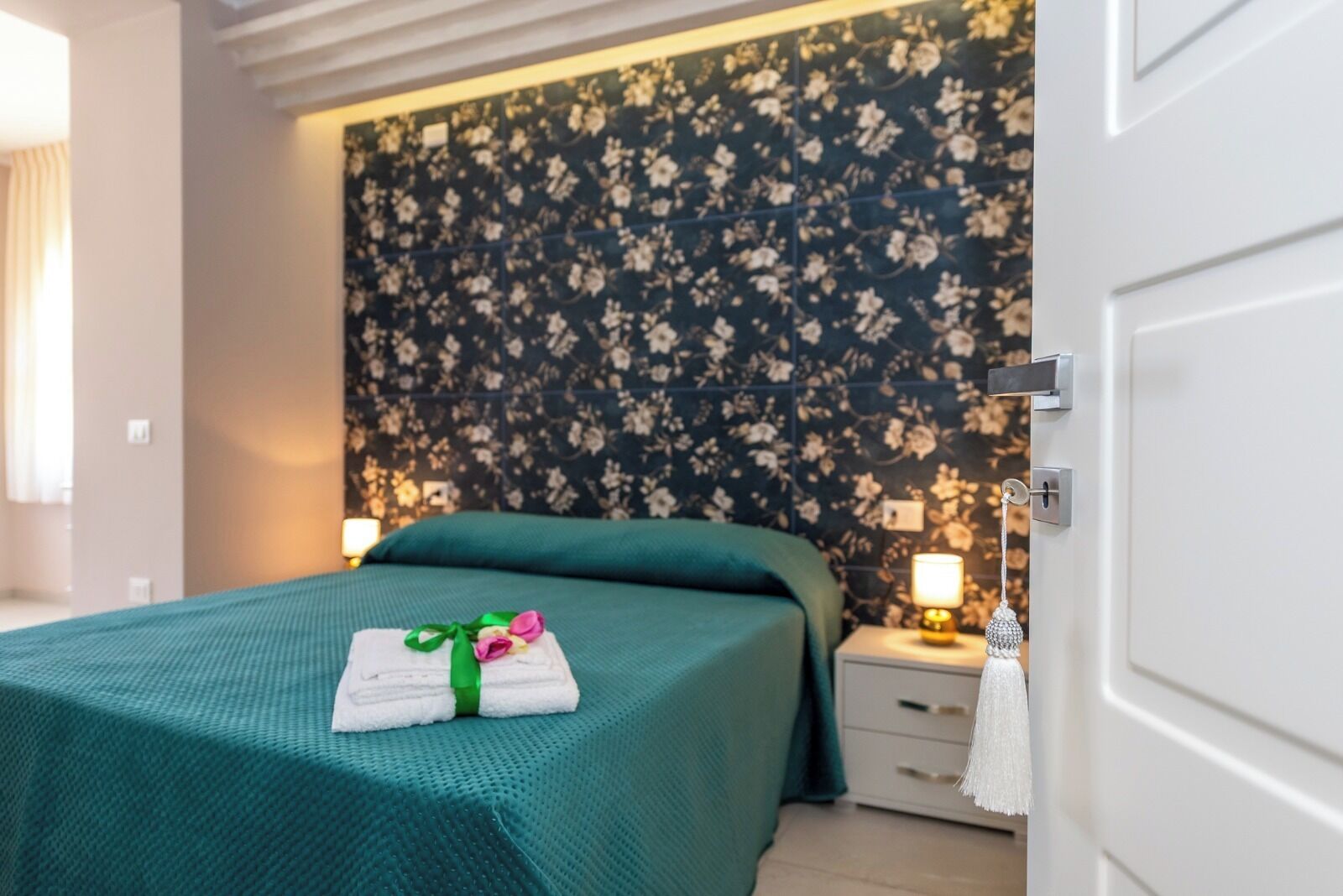 Una camera d'albergo con un letto, un comodino, una parete con carta da parati floreale e una porta.