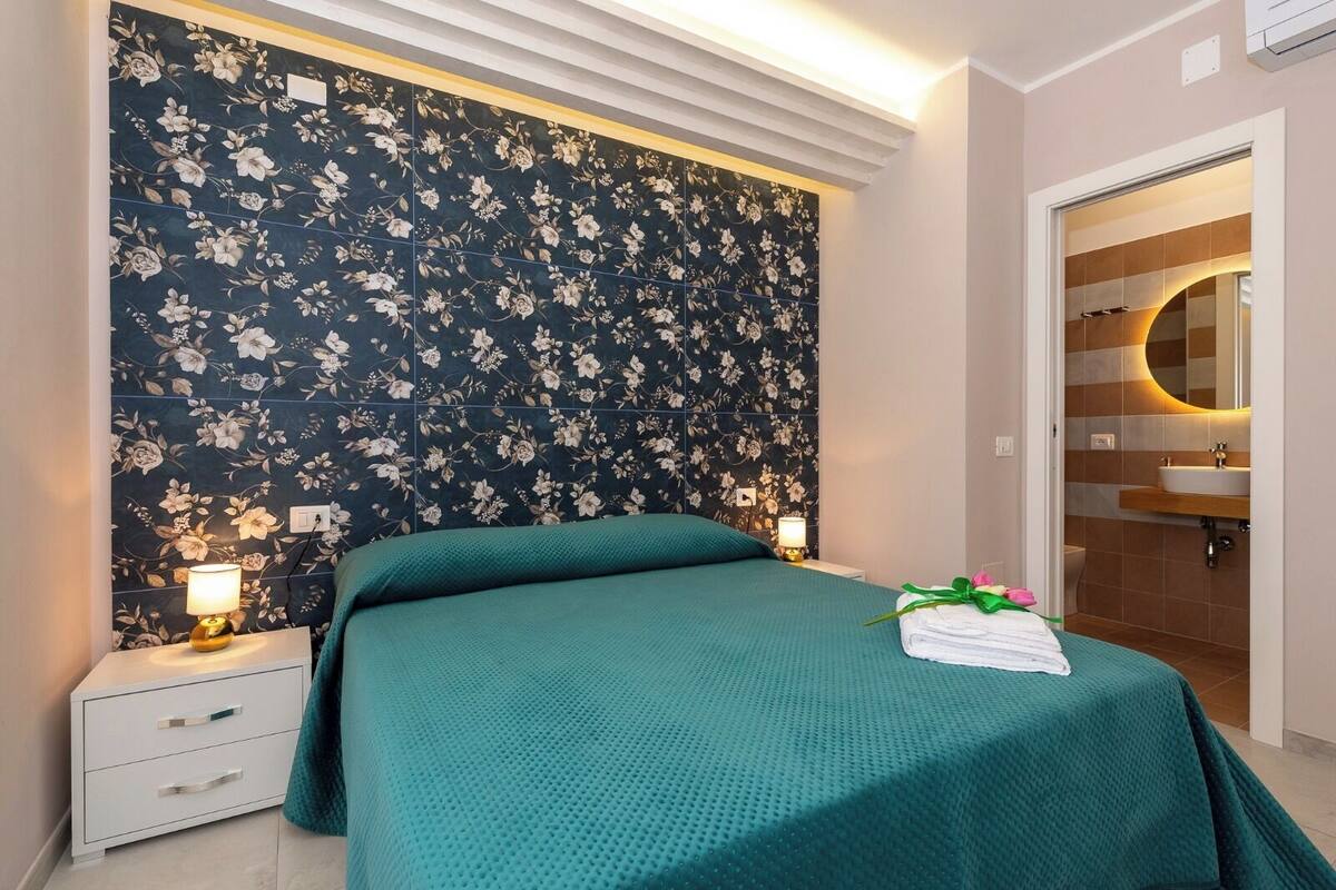 A bedroom with a floral wallpaper, a green bedspread, a bedside lamp, a nightstand, and a bathroom visible through an open door.
