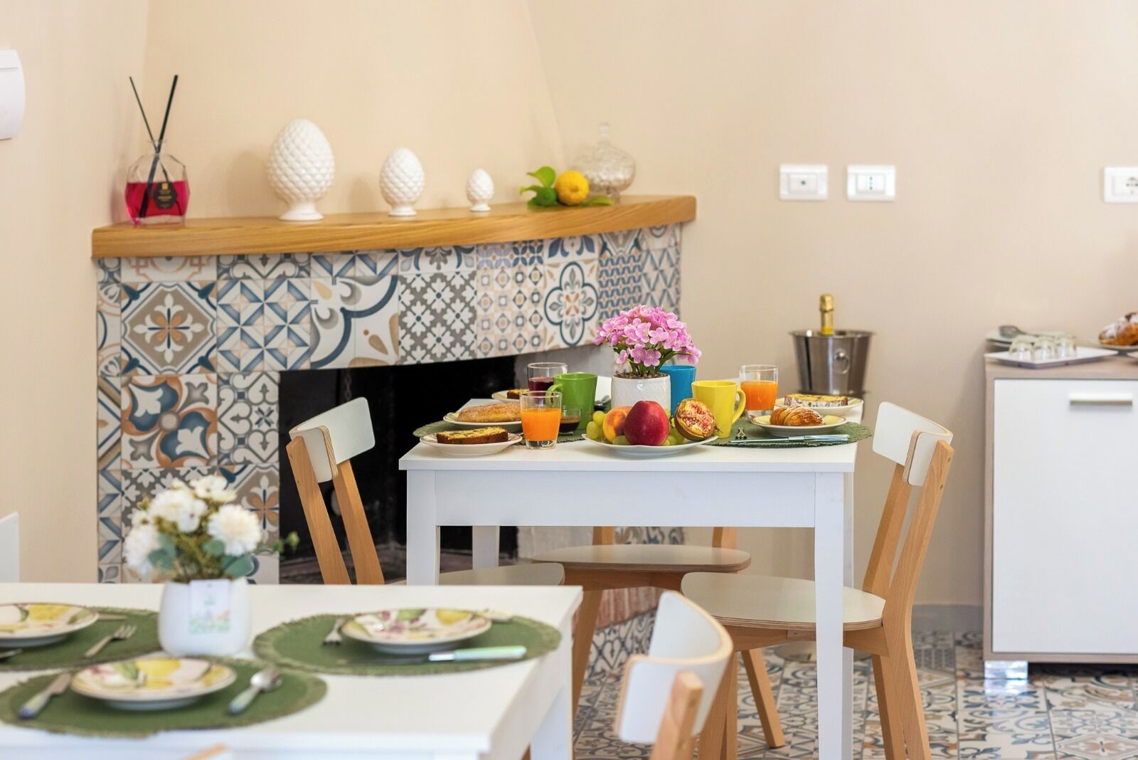 A dining area with a tiled fireplace, a table set with plates, glasses, and a fruit bowl.
