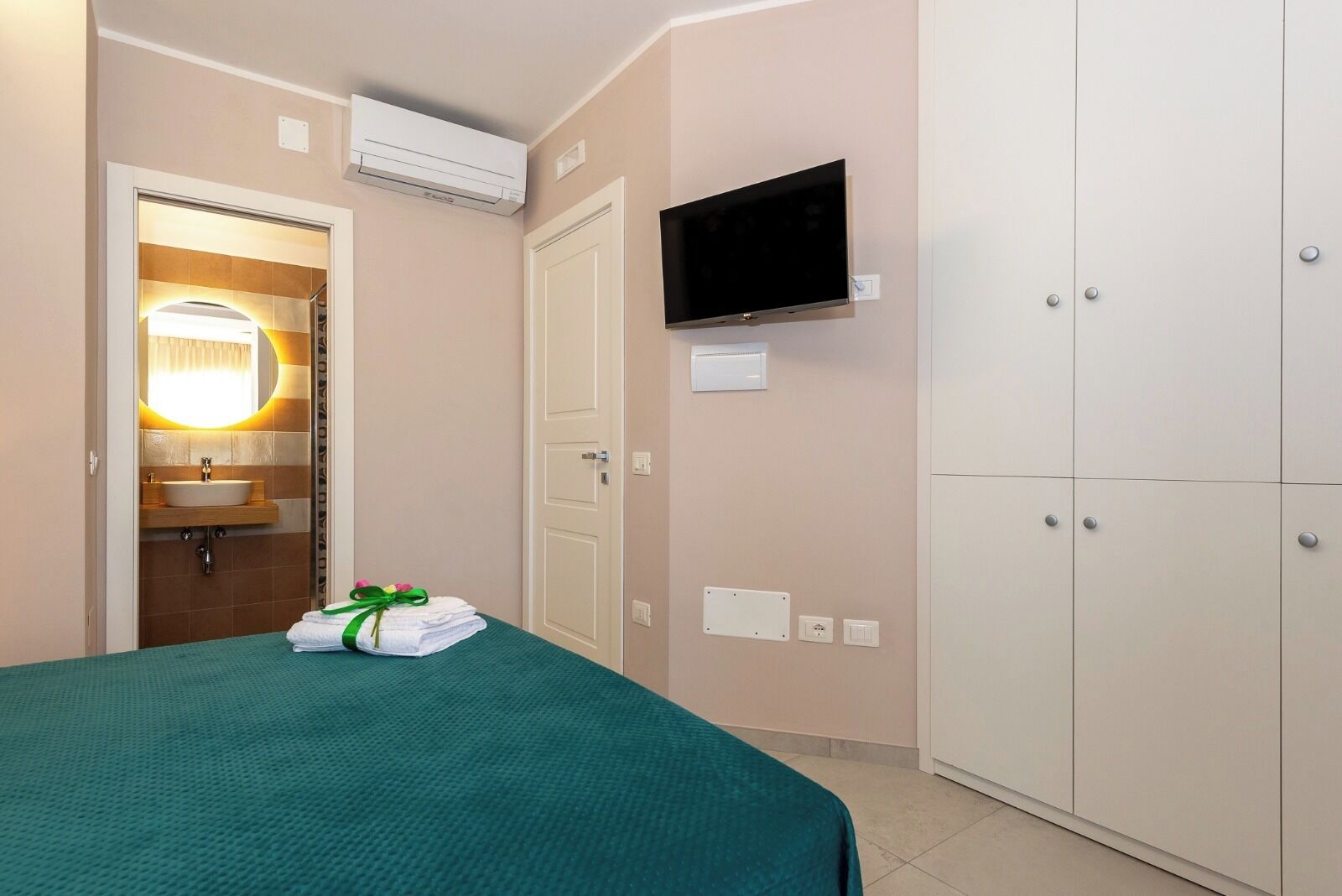 A hotel room with a bed, a TV mounted on the wall, a wardrobe, a bathroom with a sink and mirror, and an air conditioning unit.