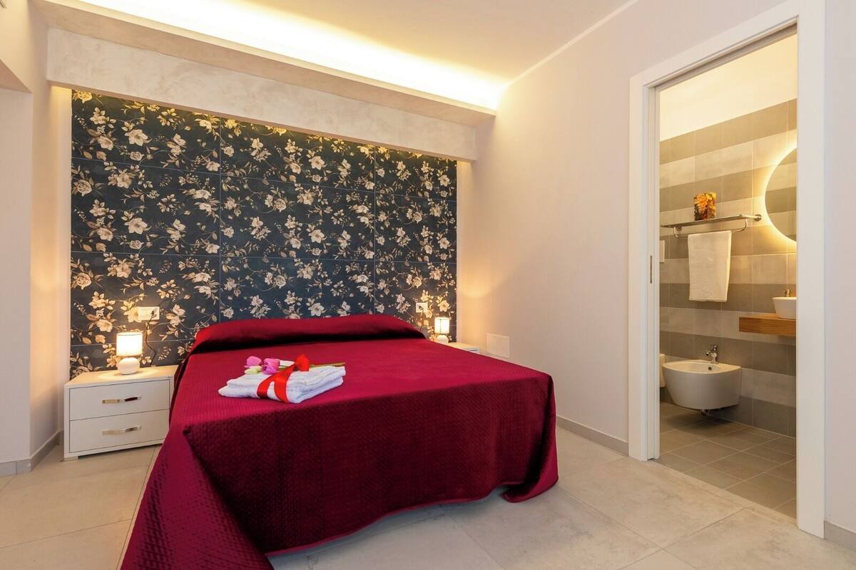 A hotel room with a bed, a nightstand, a floral wallpaper, and a bathroom visible through an open door.