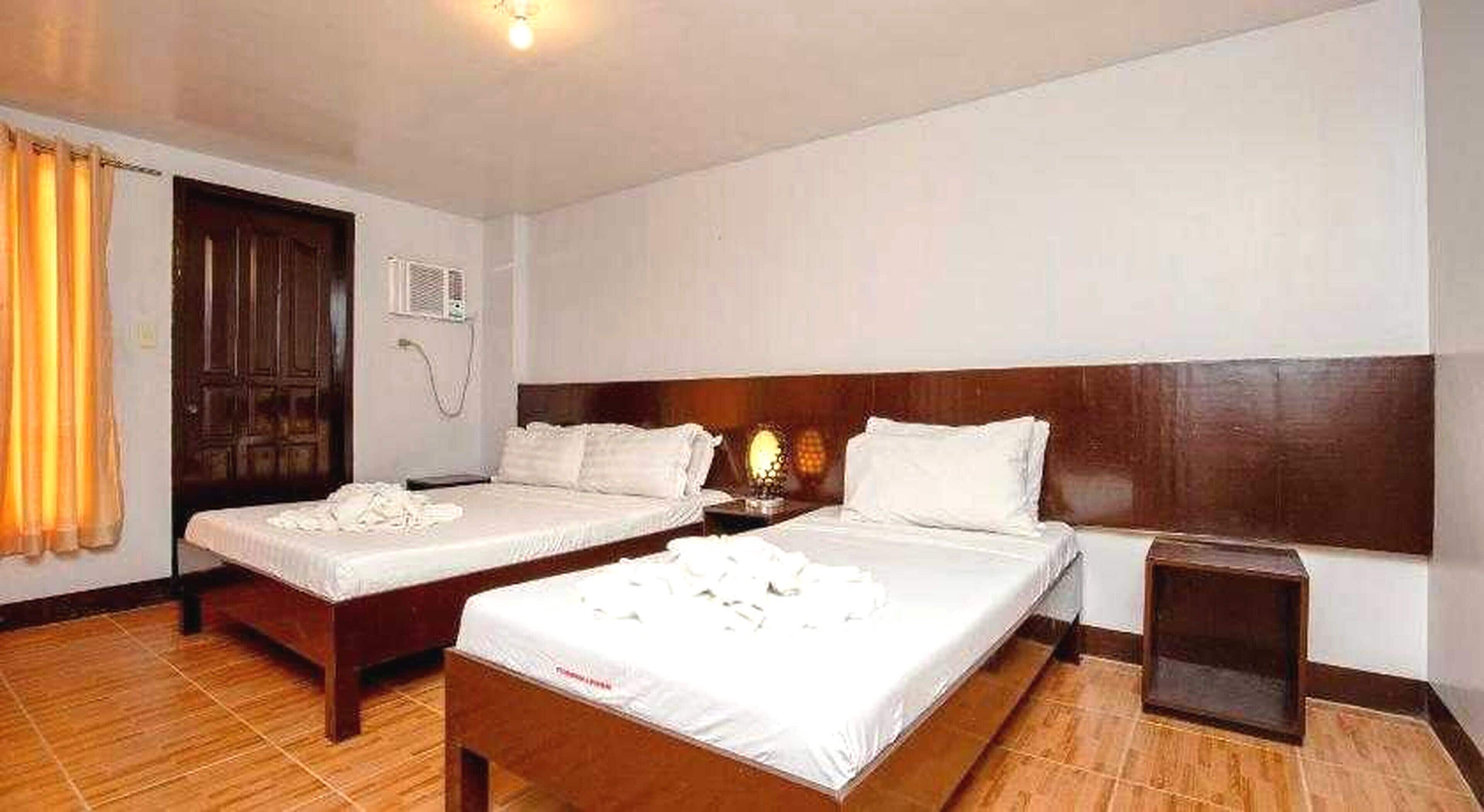 A hotel room with two beds, a wooden headboard, a bedside table, a lamp, and a window with curtains.
