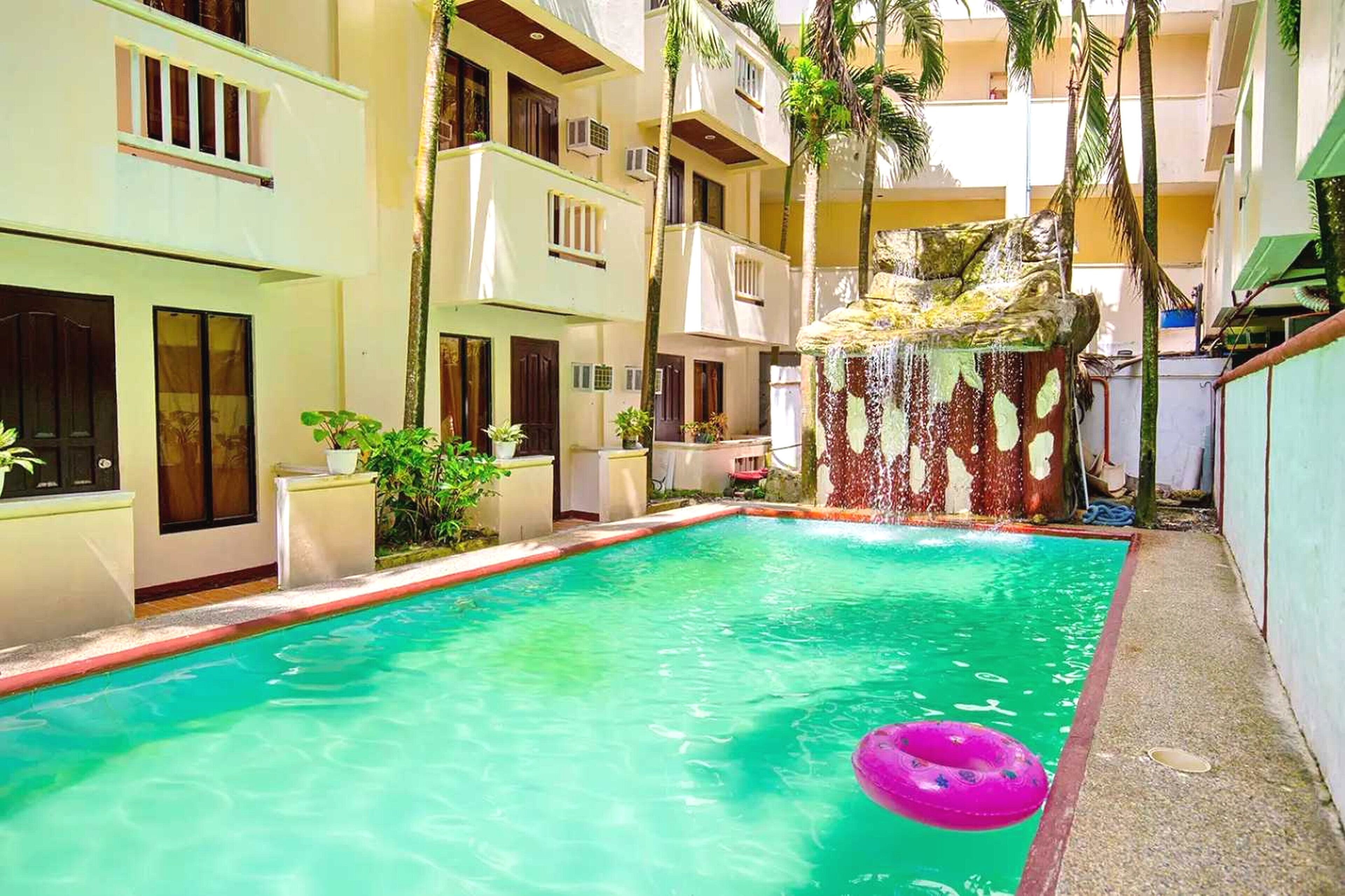 Una piscina con cascada, rodeada de edificios de varios pisos con balcones y plantas en macetas.