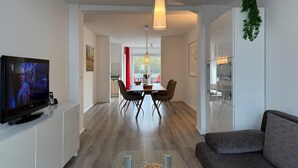 A modern living room with a flat-screen TV, a sofa, a dining area with chairs, and a kitchen with a refrigerator. - Apartment 'Traveperle 1' with Private Terrace and Wi-Fi (Lübeck)