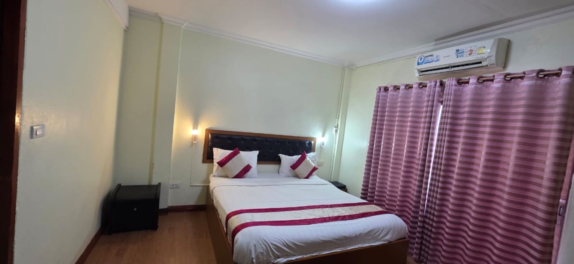 A hotel room with a bed, a bedside table, an air conditioning unit, and a curtain.