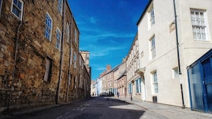 A narrow street lined with historic stone buildings and modern structures, with a clear blue sky above. - St John's College (Durham)
