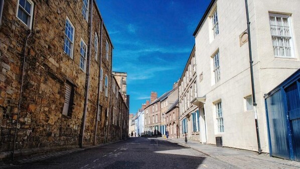 A narrow street lined with historic stone buildings and modern structures, with a clear blue sky above.