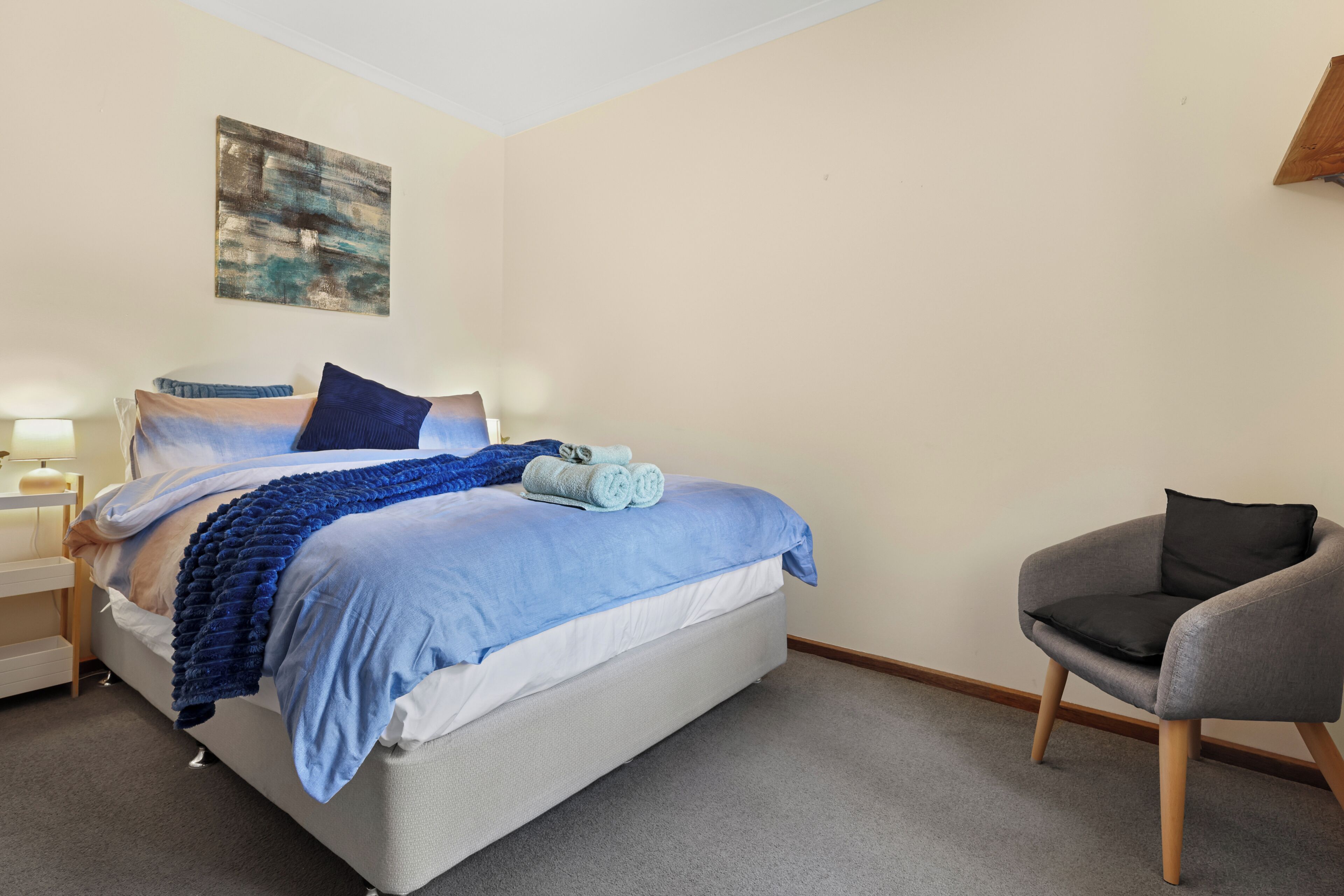 A neatly made bed with a blue blanket and pillows, a gray armchair, a bedside table with a lamp, and a painting on the wall.