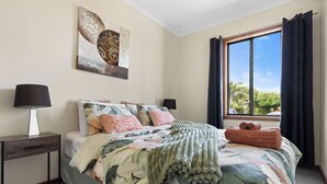 Ein Schlafzimmer mit Bett, Nachttisch, Lampe, Fenster mit Vorhängen und einem Wandbild.