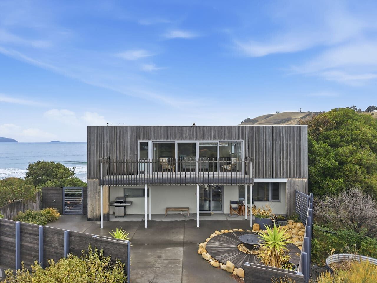 A modern two-story house with a balcony, a patio with a circular seating area, and a view of the sea.