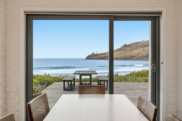 A room with a large sliding glass door leading to a deck with a table and chairs, overlooking a beach and ocean.