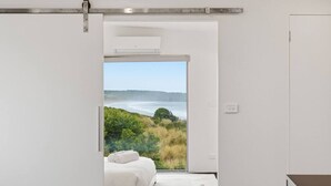 A bedroom with a bed, a wooden headboard, a white wall, and a view of a coastal landscape. - Beachfront Escape in Clifton (Clifton Beach)