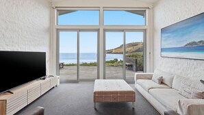 A modern living room with a large TV, a sofa, a coffee table, and a sliding glass door leading to a deck with a view of the ocean. - Beachfront Escape in Clifton (Clifton Beach)