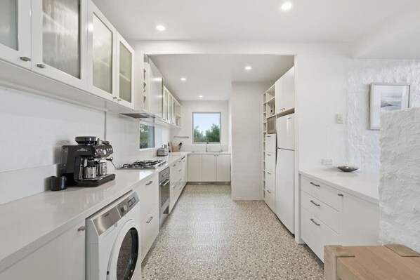 A modern kitchen with white cabinetry, a coffee machine, a washing machine, and a window providing natural light. - Beachfront Escape in Clifton (Clifton Beach)
