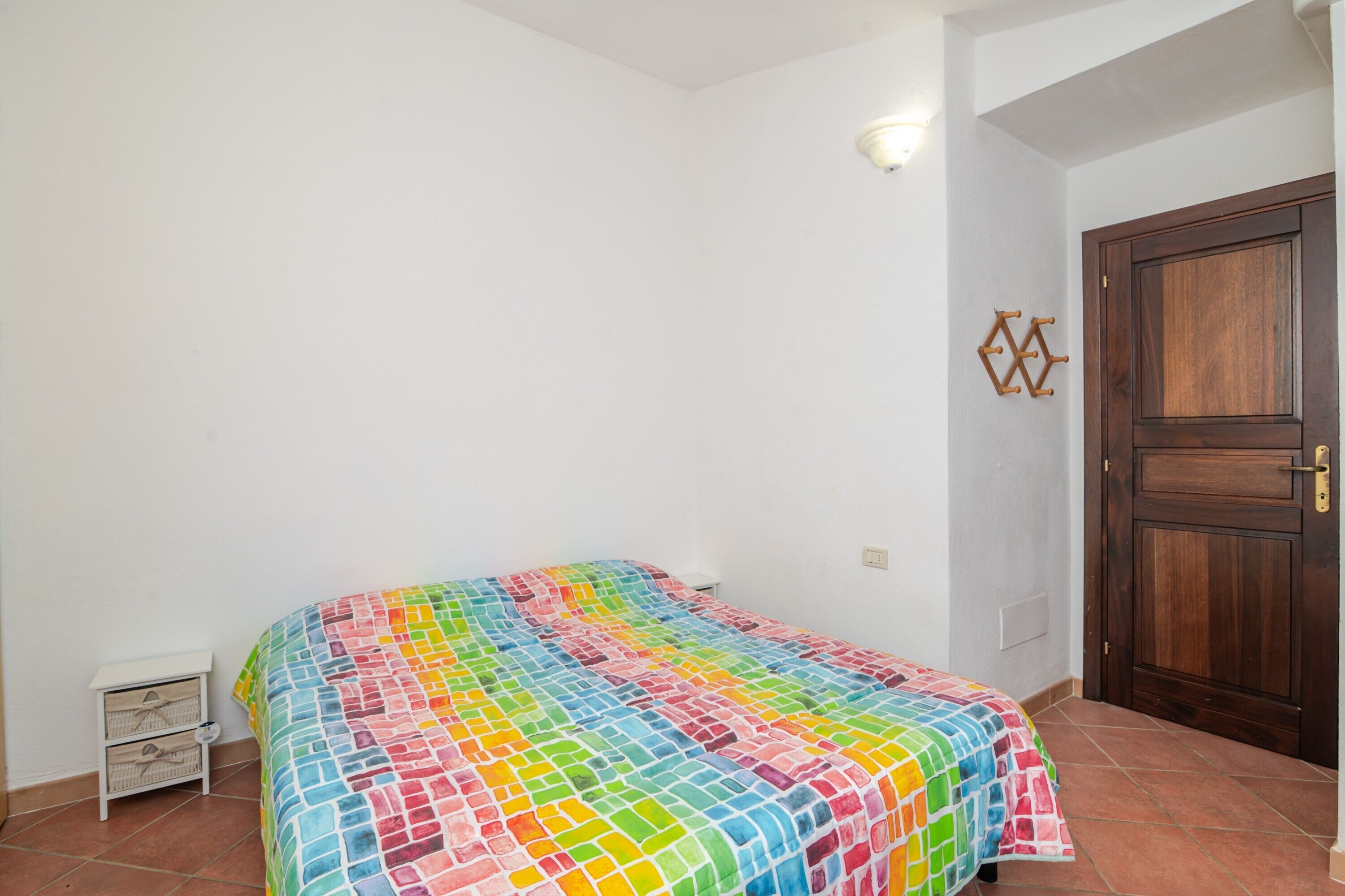 A bedroom with a colorful bedspread, a wooden door, and a small white nightstand.