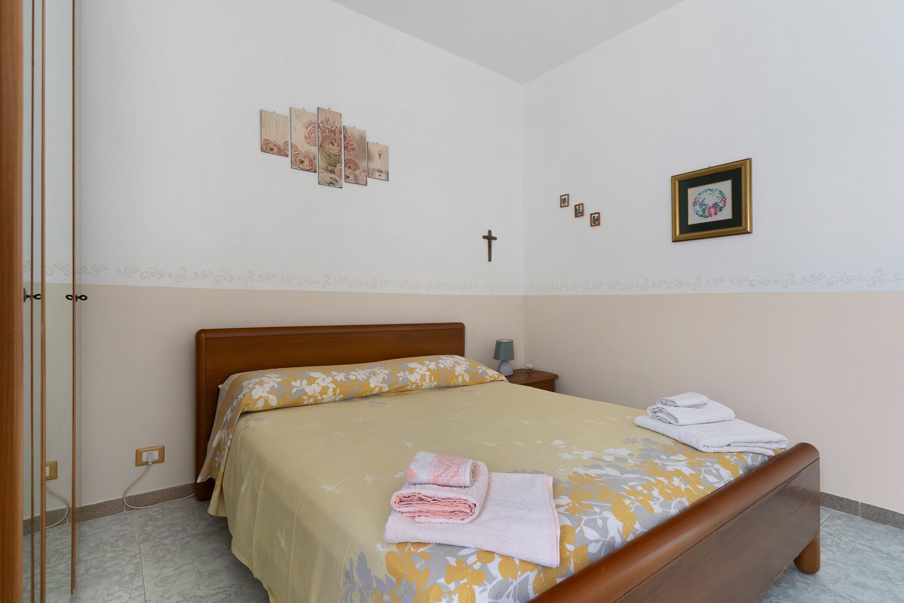 A bedroom with a bed, a nightstand, a wall-mounted cross, and a framed picture.
