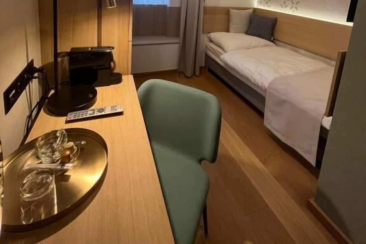 Una habitación de hotel con cama, escritorio con lámpara, silla, ventana con cortinas y pared con diseño decorativo.