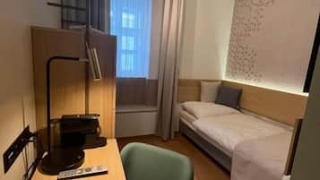 A hotel room with a bed, a desk with a lamp, a chair, a window with curtains, and a wall with a decorative pattern.