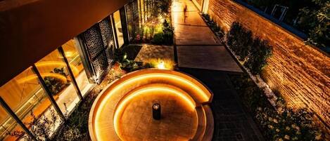 Un espacio exterior moderno con una estructura circular de madera iluminada por luces cálidas, rodeada de plantas y un muro de ladrillo.